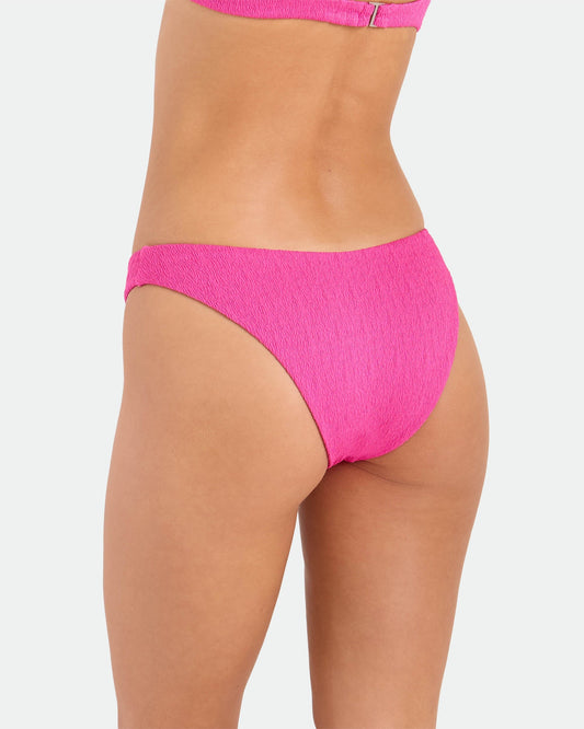 A person wears the Volcom Women's Pucker Up 90s V Bikini Bottom in bright pink, made from recycled fabric, shown from the back with part of the matching bikini top and lower back visible against a plain white background.