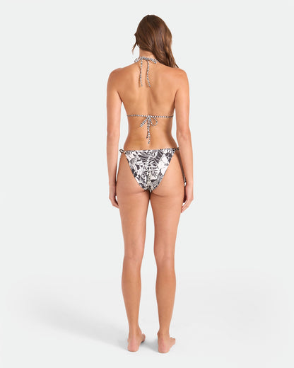 A woman stands barefoot facing away, wearing the Volcom Women's Hibiscus Diamond Reversible Triangle Top with a striped design and black-and-white floral bikini bottoms, set against a plain white background.