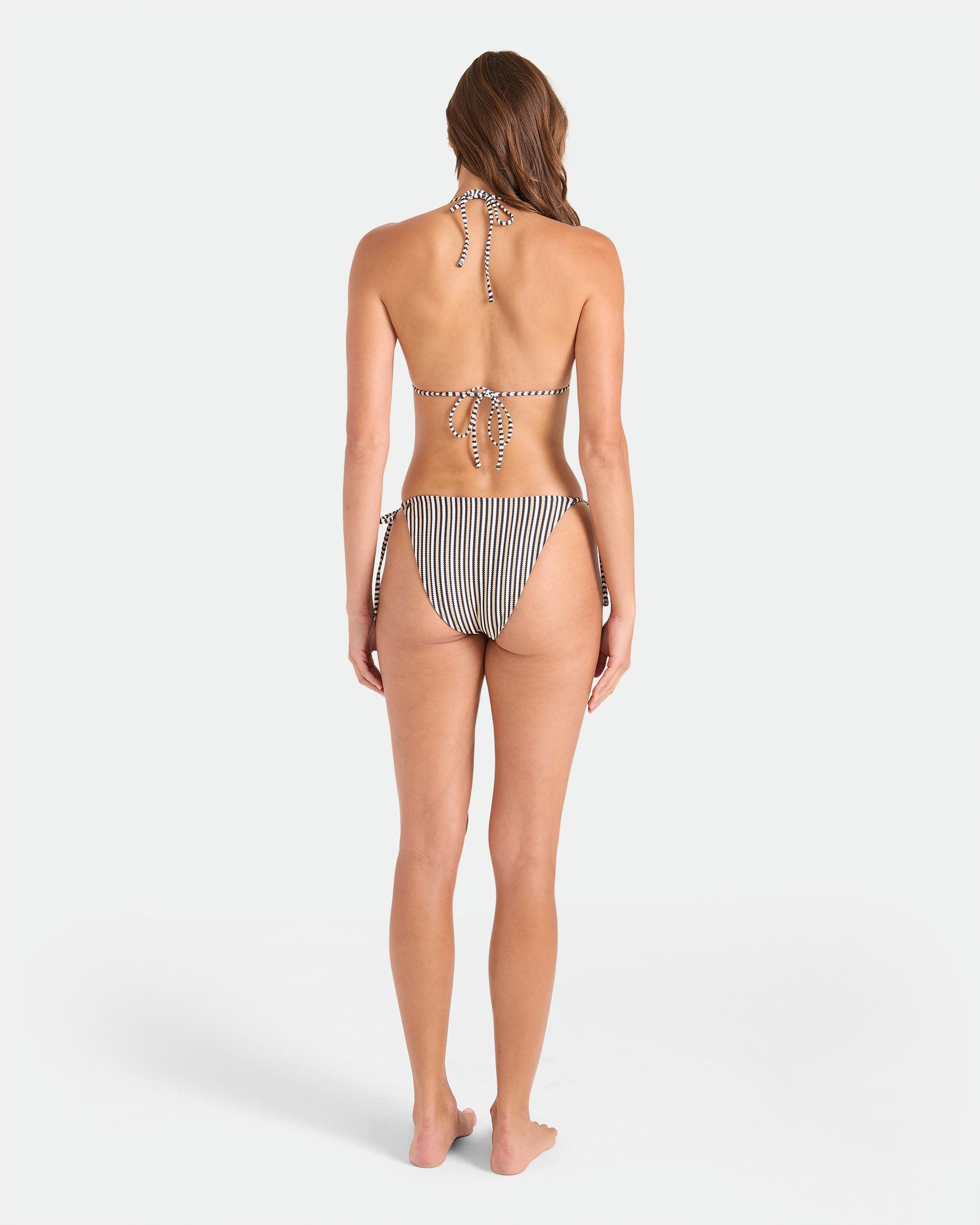 A woman stands barefoot facing away, wearing the Volcom Women's Hibiscus Diamond Reversible Triangle Top with adjustable tie straps and a black-and-white striped design, made from recycled nylon, against a white background.
