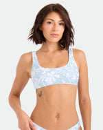 A woman with short brown hair models the Volcom Women's Tribal Stone Scoop Bralette, made from recycled nylon, in a light blue and white pattern, standing against a plain background and displaying a butterfly tattoo on her ribcage.