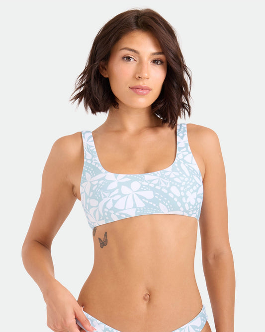 A woman with short brown hair models the Volcom Women's Tribal Stone Scoop Bralette, made from recycled nylon, in a light blue and white pattern, standing against a plain background and displaying a butterfly tattoo on her ribcage.