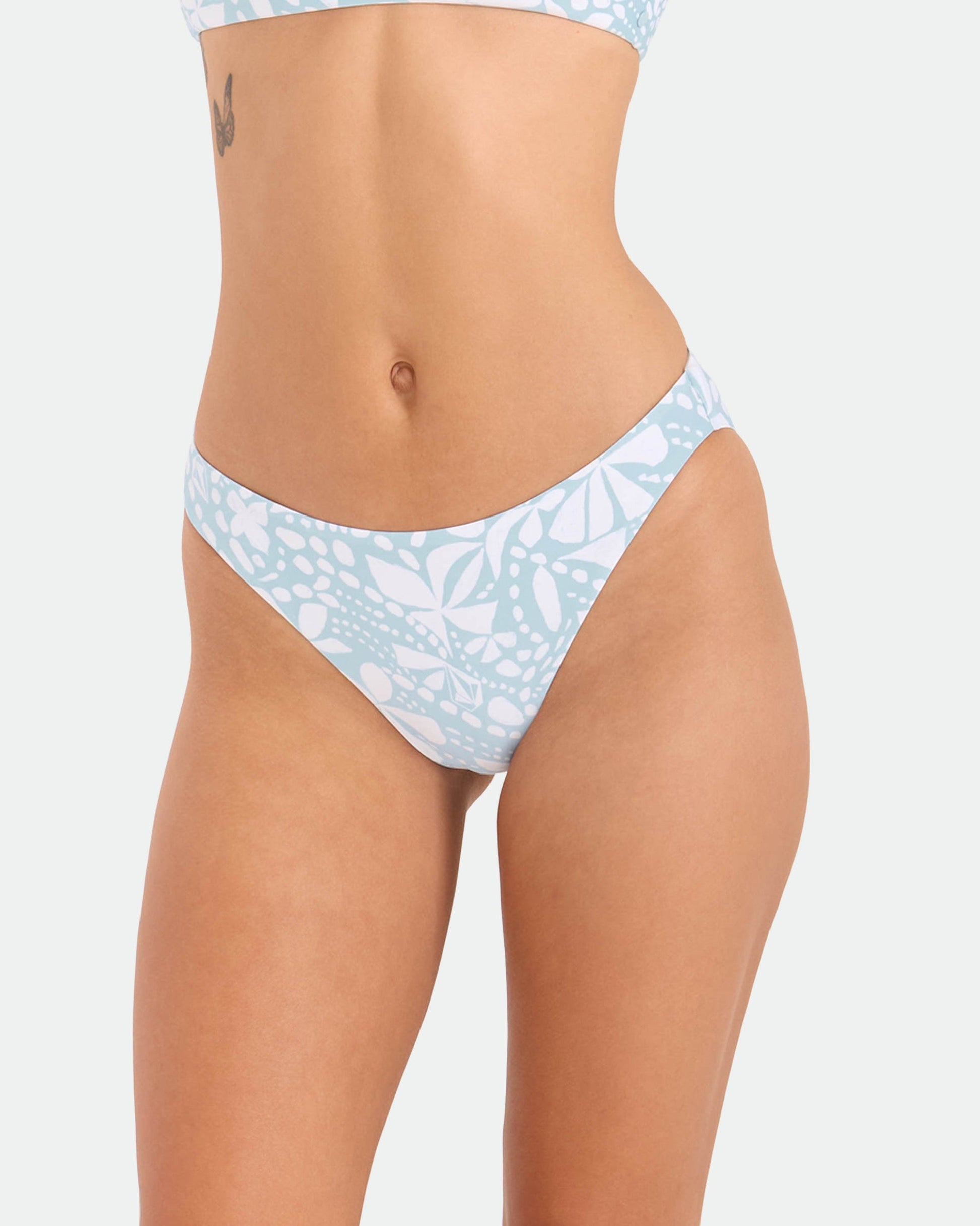 A woman models Volcom's Women's Tribal Stone Hipster Full Bikini Bottom in Summer Song, made from recycled nylon; she stands against a plain white background, showing her midsection, upper thighs, and a small tattoo near her waist.