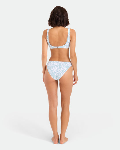 A woman stands barefoot with her back to the camera, wearing Volcom’s Women’s Tribal Stone Hipster Full Bikini Bottom in Summer Song, made from recycled nylon. The background is plain white.