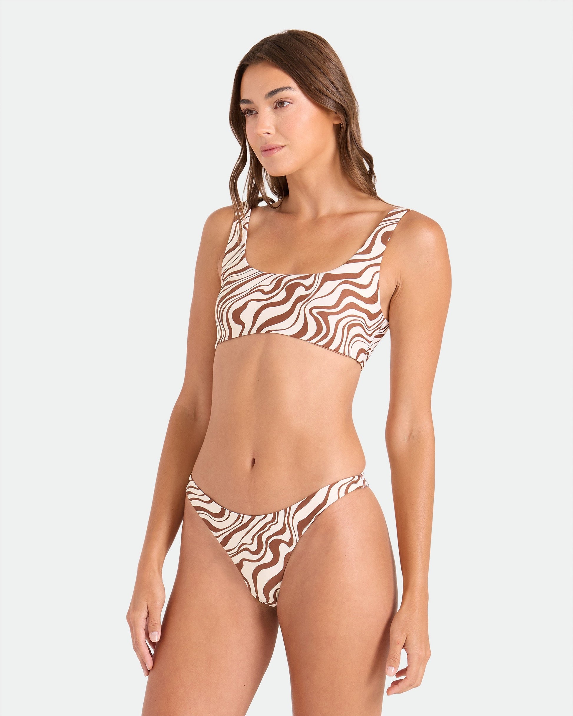 A woman poses against a white background wearing the Volcom Women's Coco Retro Bloom Scoop Bralette Reversible bikini, featuring adjustable straps, a brown and white wavy pattern, high-cut bottoms, and crafted from recycled nylon.
