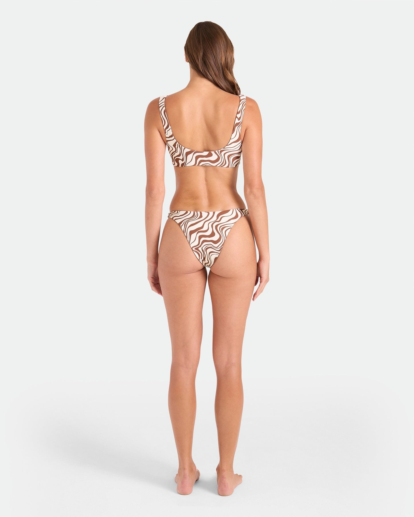 A woman with long hair poses barefoot, facing away in the Volcom Women's Coco Retro Bloom Scoop Bralette Reversible, featuring a brown and white wavy print. Made from recycled nylon with adjustable straps, set against a plain white background.