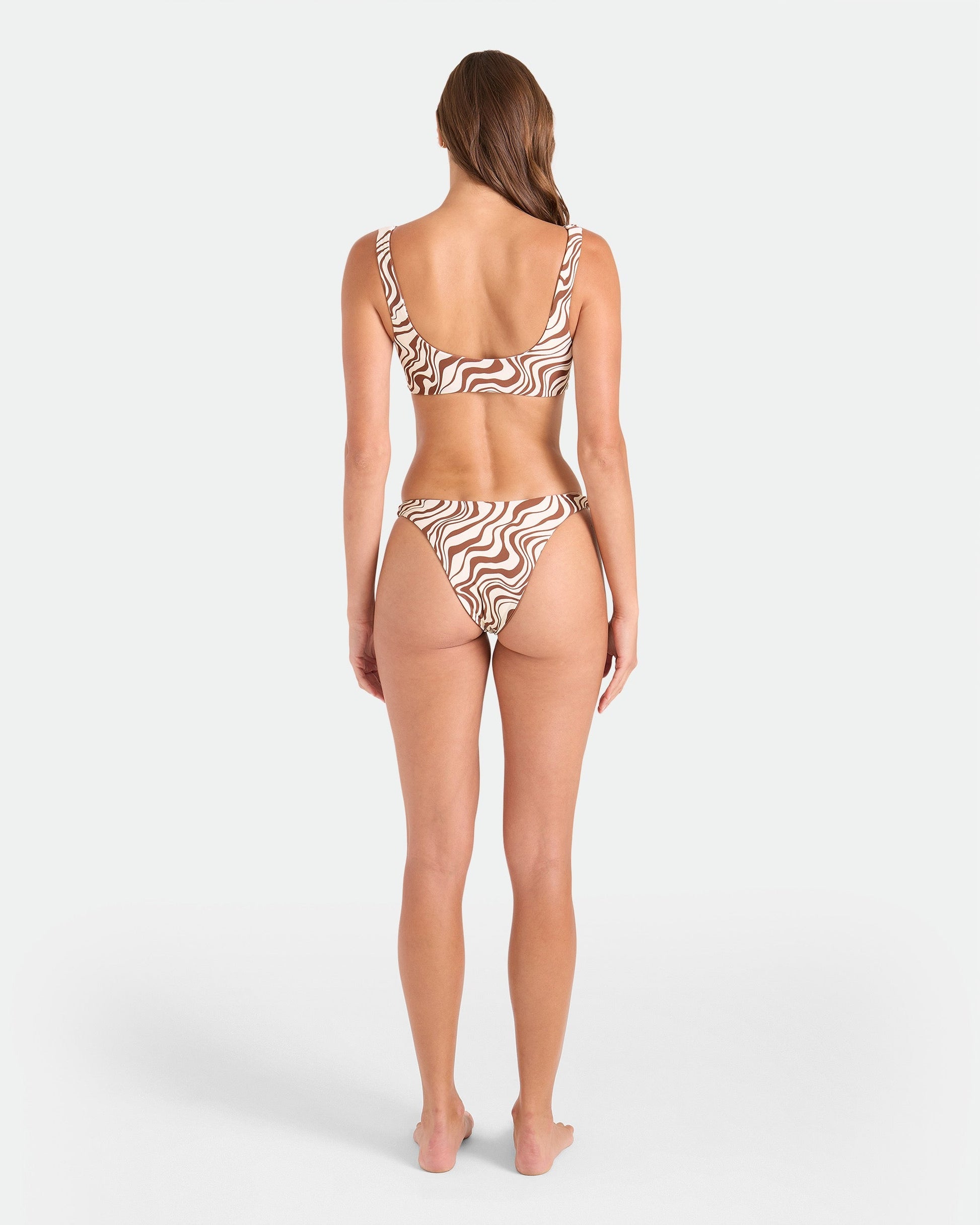 A woman with long hair poses barefoot, facing away in the Volcom Women's Coco Retro Bloom Scoop Bralette Reversible, featuring a brown and white wavy print. Made from recycled nylon with adjustable straps, set against a plain white background.
