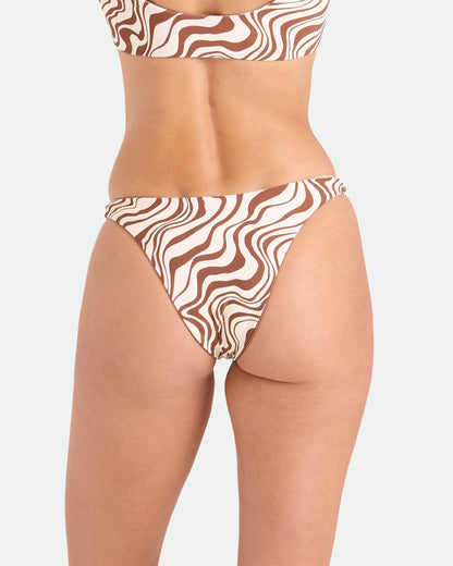 A person models the Volcom Women's Coco Retro Bloom Hipster Cheeky Reversible Bikini Bottom, featuring a brown and white wavy pattern, shown from the waist down with their back to the camera against a plain background.