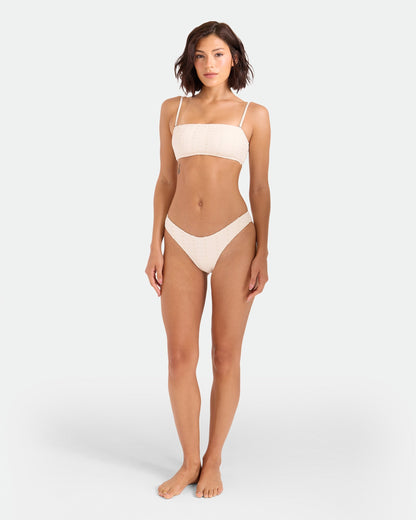 A woman with short brown hair stands barefoot against a plain light background, wearing the Volcom Women's Coco Low Tide 90s V Bottom Skimpy bikini, featuring a light-colored strapless top and matching high-leg cut bottoms.