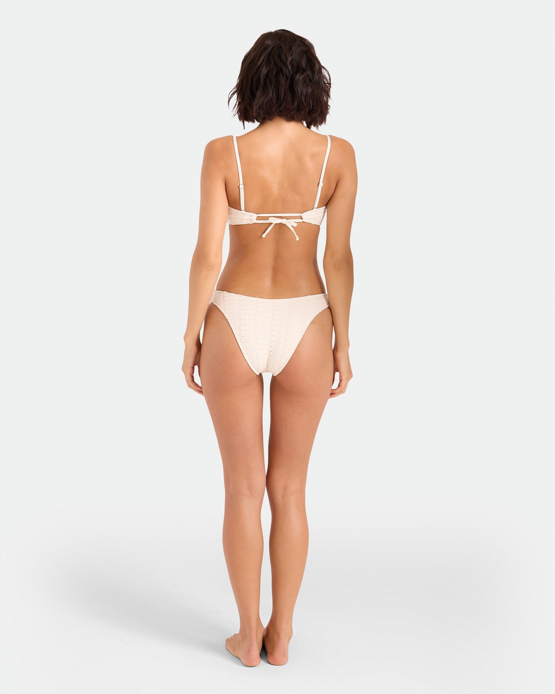 A woman stands barefoot facing away, wearing the Volcom Women's Coco Low Tide 90s V Bottom Skimpy bikini, featuring a tied back top and minimal coverage, set against a plain white background.