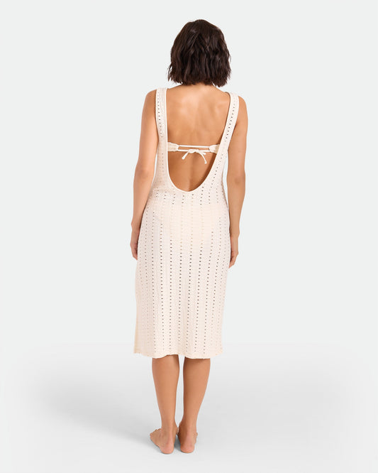 A woman stands barefoot facing away, wearing the Volcom Women's Coco Low Tide Midi Dress—a sleeveless, open-back knit body-con tank dress in light cream with a tie strap and below-the-knee length, set against a plain white background.