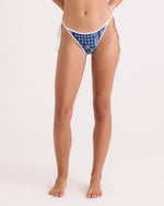 A woman stands barefoot against a plain light background wearing Volcom's Women's Bandana Tie Side Teeny Reversible bikini bottoms, featuring a blue and white pattern and crafted from recycled swimwear materials. Her head is not visible in the image.