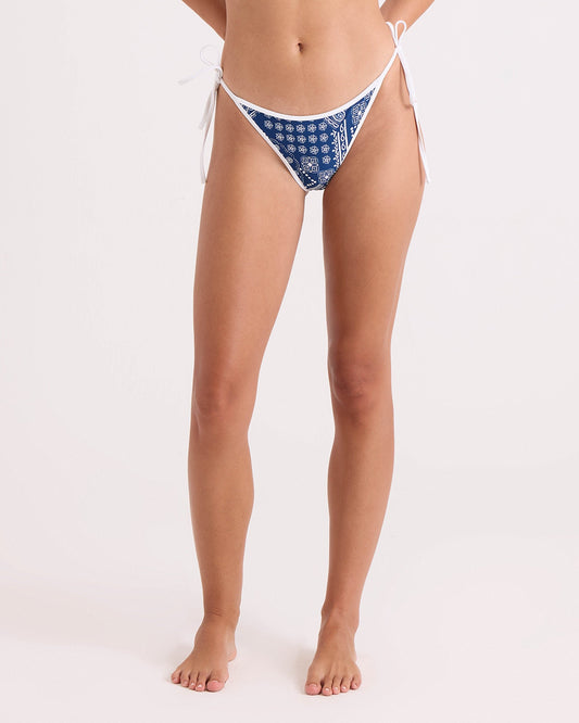 A woman stands barefoot against a plain light background wearing Volcom's Women's Bandana Tie Side Teeny Reversible bikini bottoms, featuring a blue and white pattern and crafted from recycled swimwear materials. Her head is not visible in the image.
