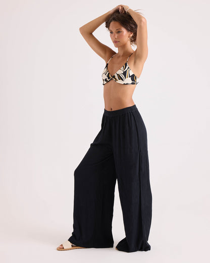 Wearing the Volcom Women's Wild Bloom Twist Bralette, a woman stands barefoot on one foot in loose black pants and white sandals, raising her arms to adjust her hair against a plain light background.