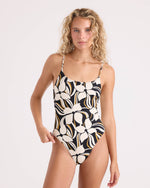 A woman with wavy blonde hair models the Volcom Women's Wild Bloom Skimpy One Piece, a black lingerie-style swimsuit featuring a striking white and beige floral print and adjustable straps, standing against a plain light background.
