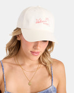 A woman wears Volcom's Women's Stone Rising Hat, a cream cotton twill baseball cap with pink text and a palm tree, featuring an adjustable strap. She pairs it with a blue checkered tank top and layered gold necklaces against a light background.