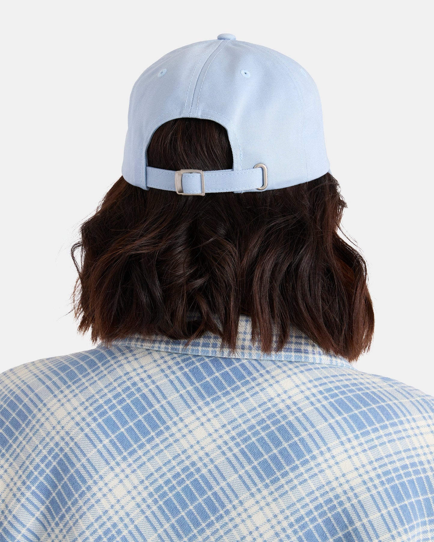 A woman with shoulder-length dark hair is seen from behind against a plain background, wearing Volcom’s Women’s Circle Stone Dad Hat in light blue and a blue-and-white plaid shirt.