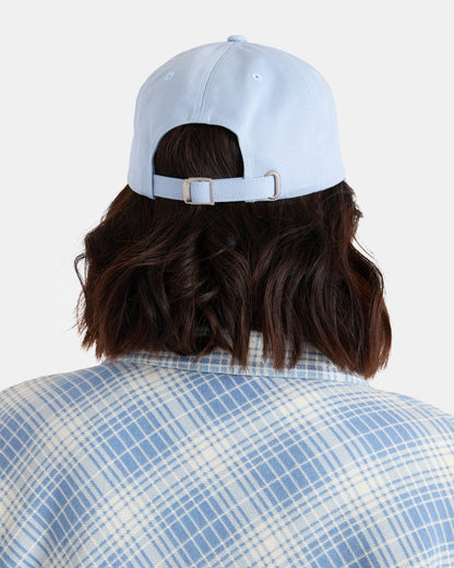 A woman with shoulder-length dark hair is seen from behind against a plain background, wearing Volcom’s Women’s Circle Stone Dad Hat in light blue and a blue-and-white plaid shirt.