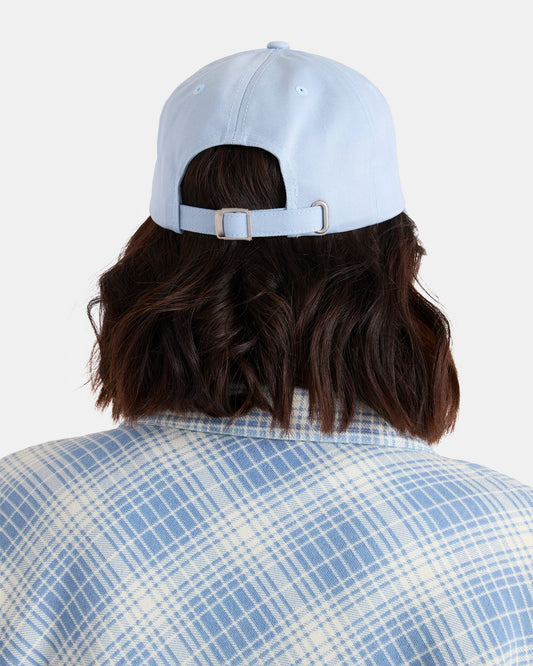 A woman with shoulder-length dark hair is seen from behind against a plain background, wearing Volcom’s Women’s Circle Stone Dad Hat in light blue and a blue-and-white plaid shirt.