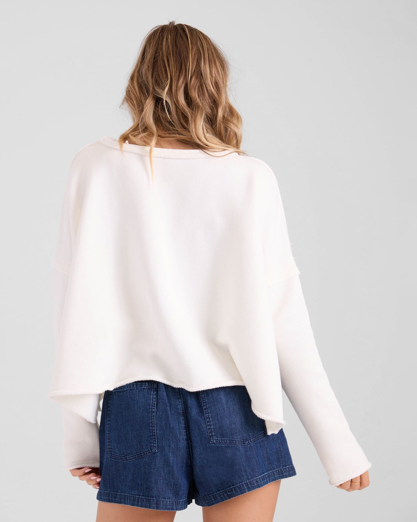 A person with wavy, light brown hair is seen from the back, wearing the Volcom Women's Gnarly relaxed fit sweatshirt paired with high-waisted dark denim shorts against a plain light background.