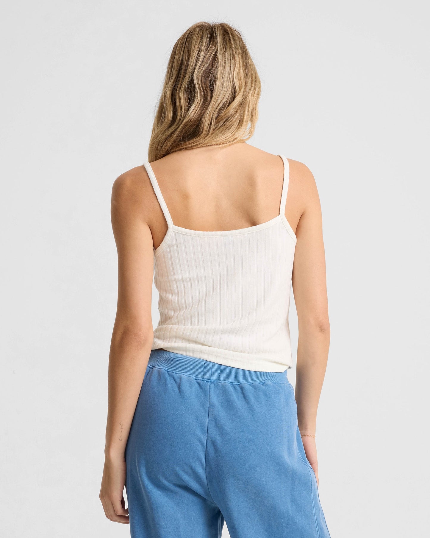 A woman with long blonde hair faces away, wearing the Volcom Women's Lived In Rib Tank and loose blue pants against a plain light gray background.