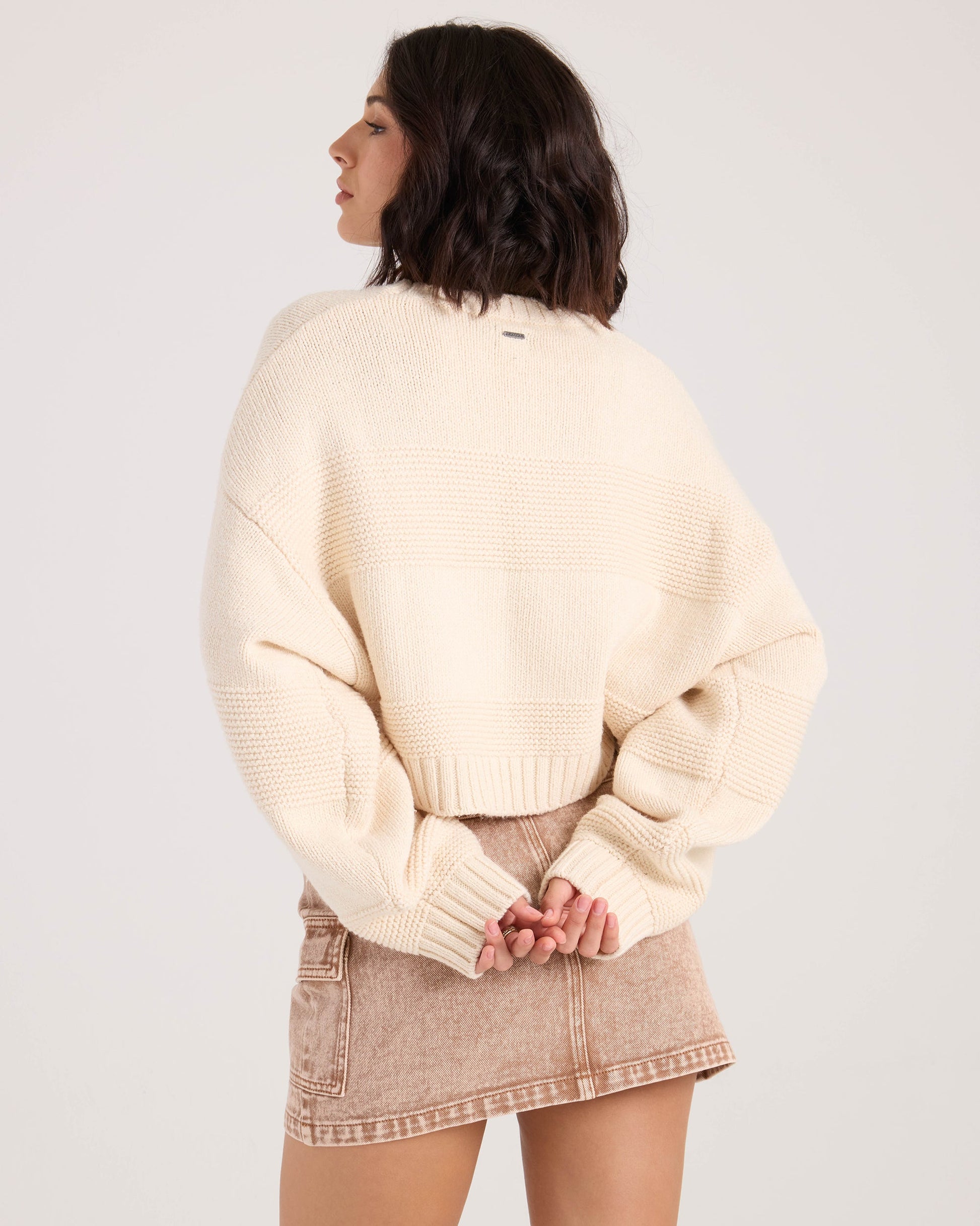 A woman with shoulder-length dark hair models the Volcom Women's Dayz Like This Sweater in white, standing with her back to the camera and hands clasped behind her.