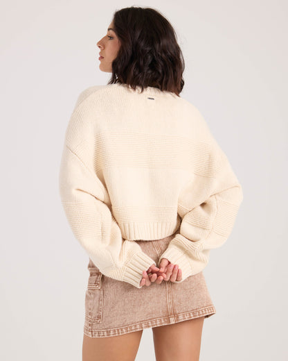A woman with shoulder-length dark hair models the Volcom Women's Dayz Like This Sweater in white, standing with her back to the camera and hands clasped behind her.