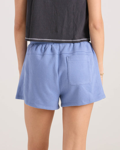 A person shown from the waist down, facing away, wears a black cropped top and light blue Volcom Women's Lived In Fleece Shorts with an elastic waistband and back pocket.