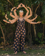 A woman stands barefoot outdoors in the Volcom Women's Midnite Ramble Dress. Behind her, people extend their arms so she appears to have many arms like a Hindu deity, with lush greenery all around.