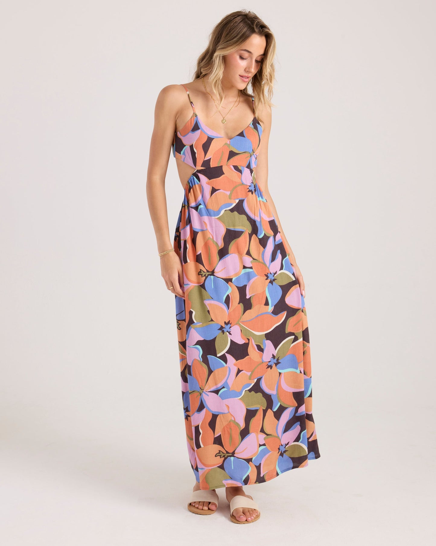 A woman with light brown hair wears the Volcom Women's Post Punk Dress, an A-line maxi with a bold floral print in orange, purple, pink, and green. She pairs it with sandals and stands against a plain white background.
