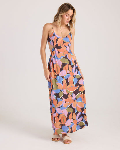 A woman with light brown hair wears the Volcom Women's Post Punk Dress, an A-line maxi with a bold floral print in orange, purple, pink, and green. She pairs it with sandals and stands against a plain white background.