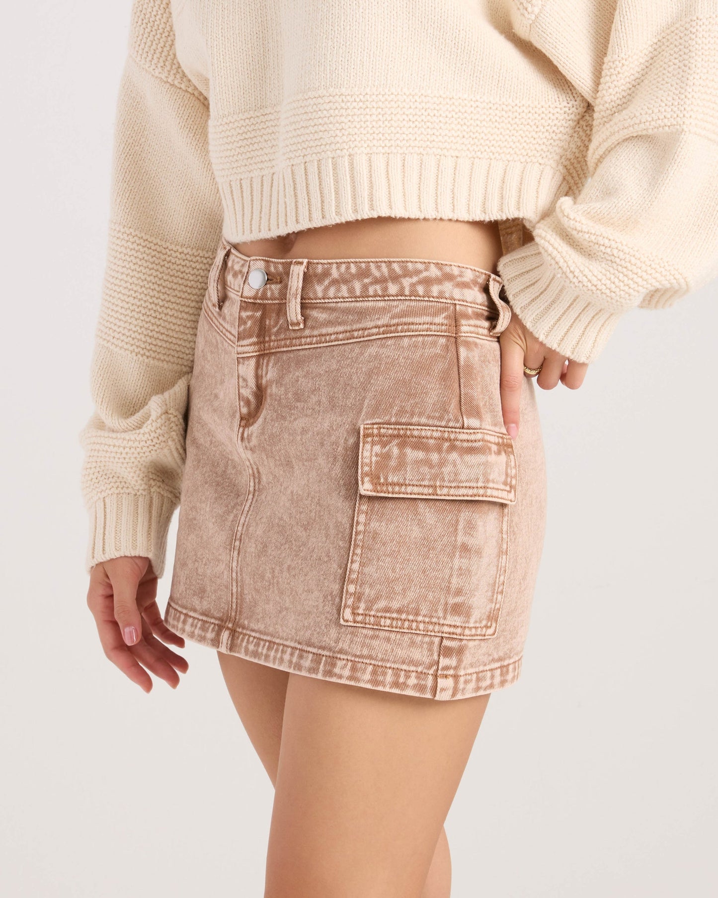 A woman shown from the waist down wears a cream knit sweater paired with the Volcom Women's Delia Cargo Mini Skirt in light brown acid wash, featuring a large side pocket.