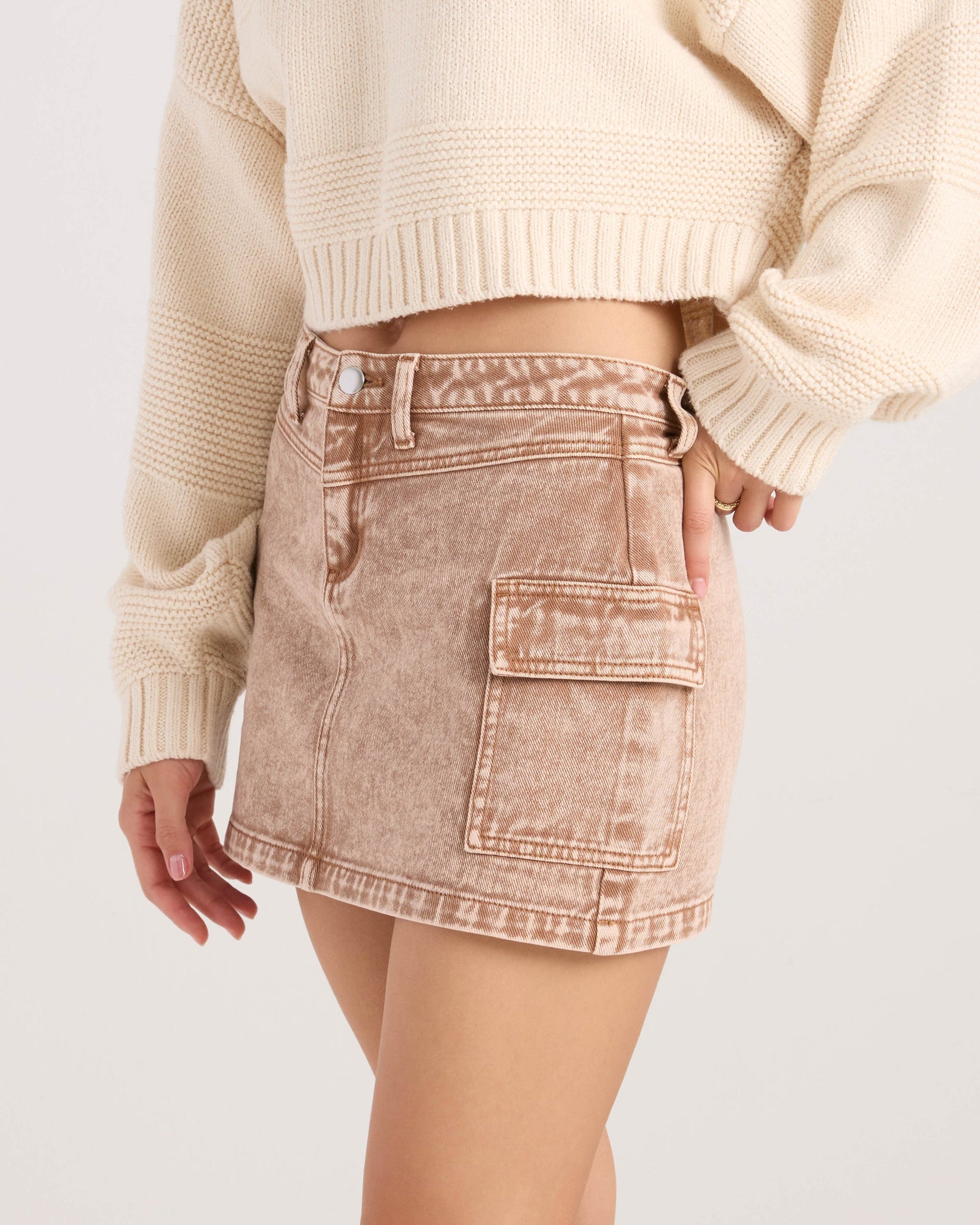 A woman shown from the waist down wears a cream knit sweater paired with the Volcom Women's Delia Cargo Mini Skirt in light brown acid wash, featuring a large side pocket.