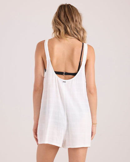 A woman with wavy blonde hair, seen from the back, wears the Volcom Women's Lived In Short Romper in white over a black bra. This lightweight cotton romper has a low scoop back, relaxed fit, and is shown against a plain light background.