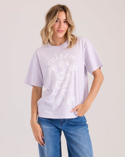 A woman with wavy blonde hair wears the Volcom Women's Burnout BF Tee—a loose lavender shirt with a white graphic—paired with blue jeans, standing against a light background looking relaxed with one hand in her pocket.