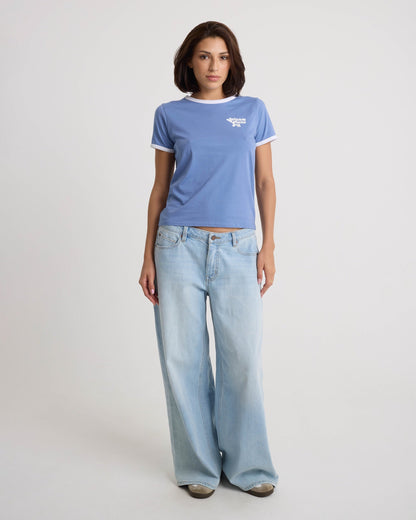 A woman faces forward with a neutral expression, wearing the Volcom Women's Brk Up Ringer Tee in Horizon Blue, paired with loose light blue jeans and sneakers against a plain white background.