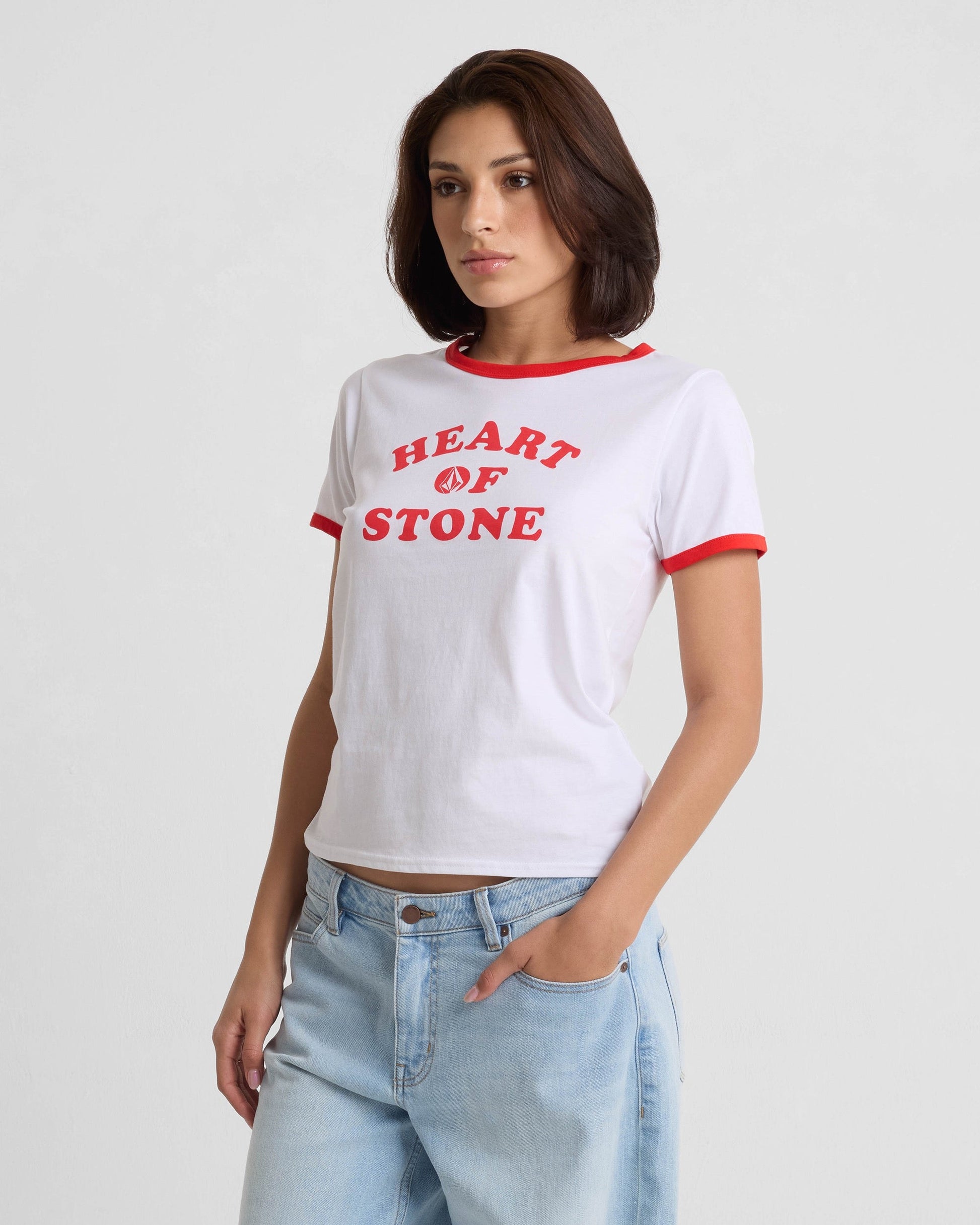 A woman with short brown hair wears the Volcom Women's Brk Up Ringer Tee—white with red trim and bold "HEART OF STONE" text—paired with light blue jeans, standing against a plain light background.
