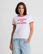 A woman with straight brown hair wears the Volcom Women's Brk Up Ringer Tee, a fitted top with red trim and bold HEART OF STONE lettering, paired with light blue jeans. She stands against a plain background with one hand in her pocket.