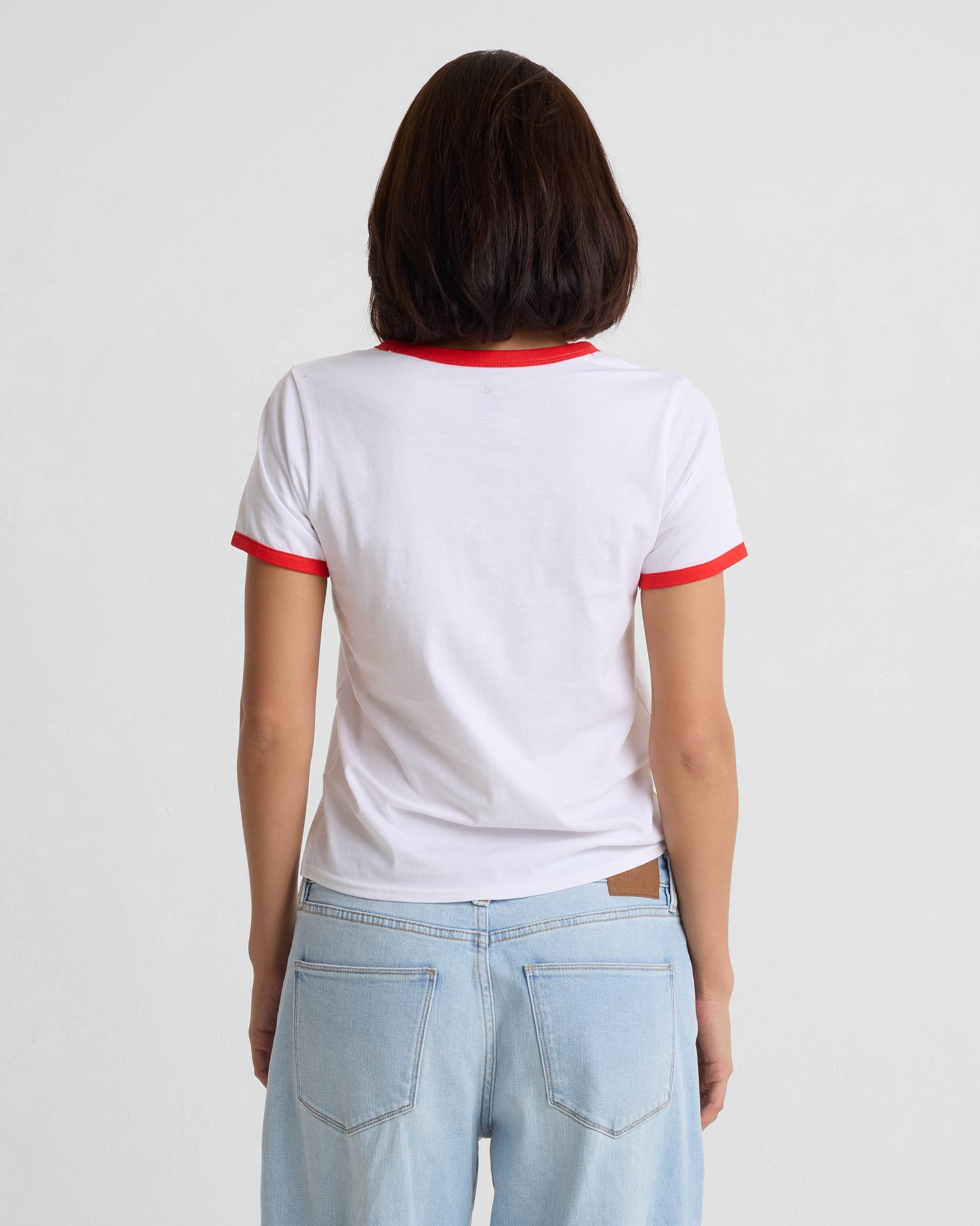 A person with medium-length brown hair, seen from behind, wears the Volcom Women's Brk Up Ringer Tee—featuring red trim on the sleeves and collar—paired with light blue jeans against a plain light background.