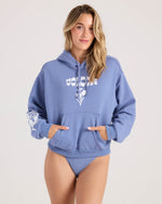 A woman with long blonde hair models the Volcom Women's 1991 Mini Hoodie in Horizon Blue with white graphic text and a front pocket, paired with matching blue bikini bottoms, against a plain light backdrop.
