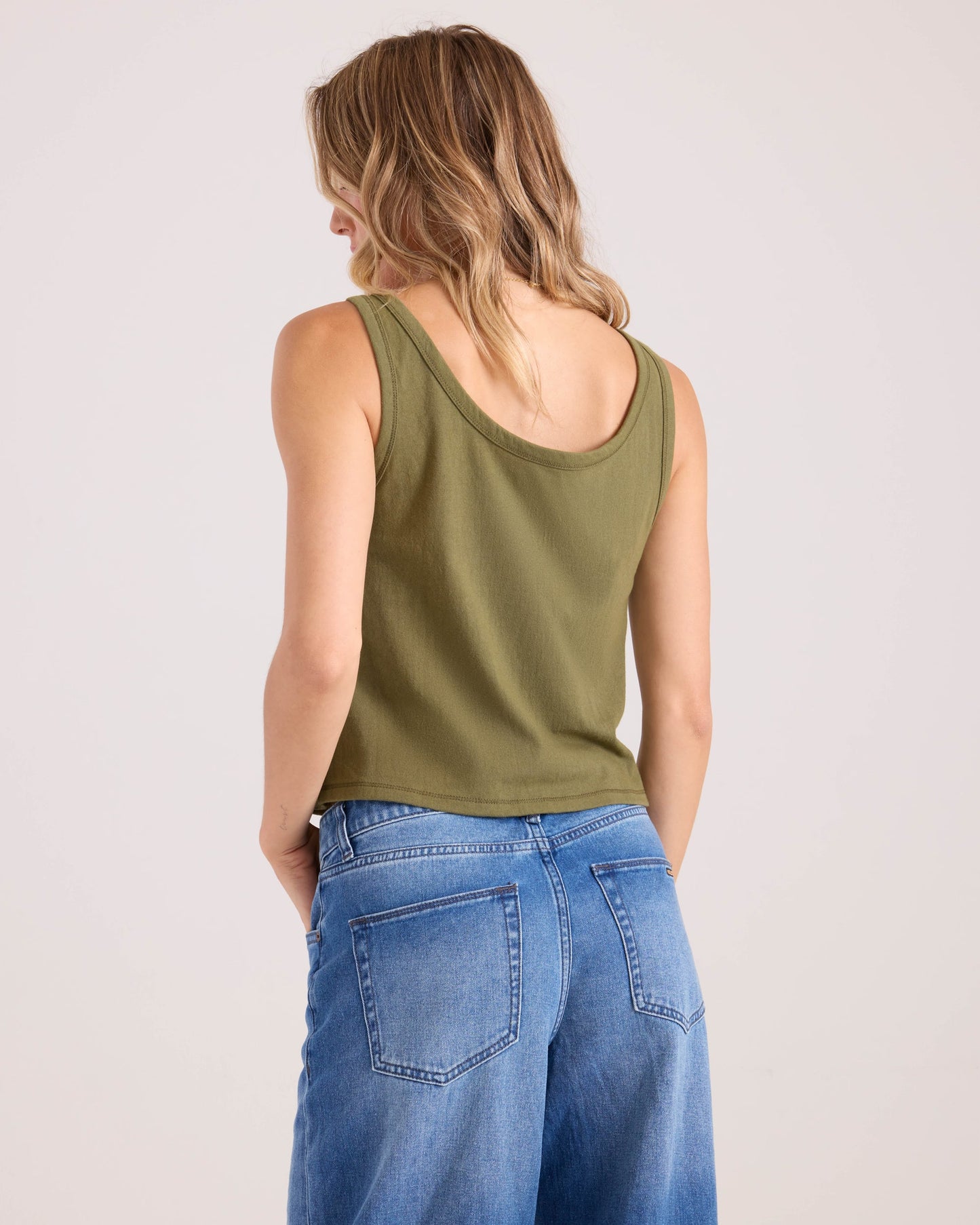 A woman with wavy, shoulder-length blonde hair is seen from behind wearing the Volcom Women's 91 Tank in olive green and high-waisted blue jeans against a plain light background.