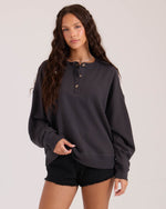 A young woman with long, wavy brown hair wears the Volcom Women's Lived In Fleece Henley and black denim shorts, standing against a plain light background and looking at the camera.
