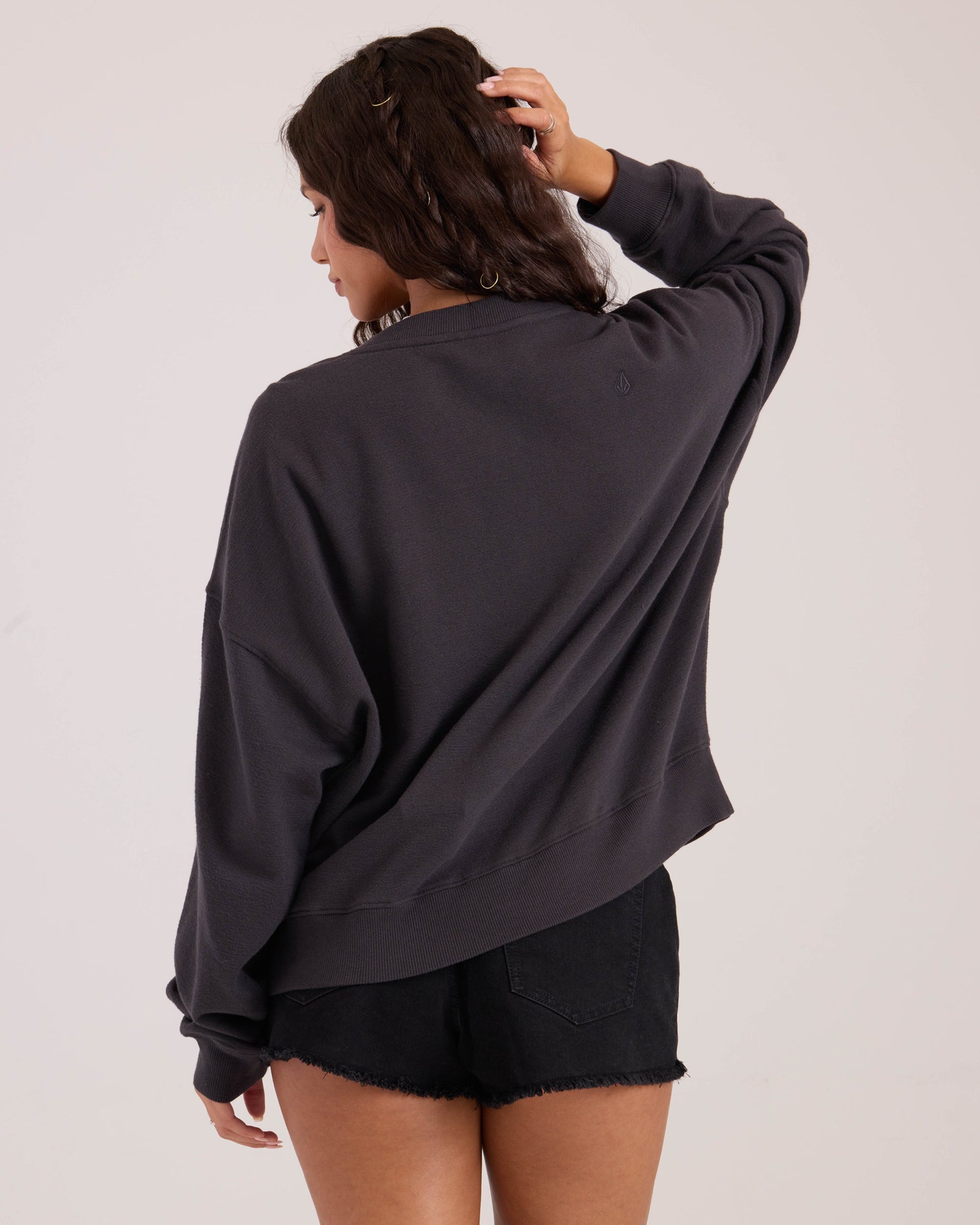 A woman with wavy brown hair wears the Volcom Women's Lived In Fleece Henley and black denim shorts, standing with her back to the camera, one hand on her head, against a plain light background.