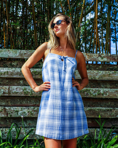 A woman poses outdoors on a sunny day wearing Volcom's Women's Folkin Stone Dress—an A-line mini with a blue plaid pattern—and sunglasses, standing in front of a wooden fence and tall bamboo.