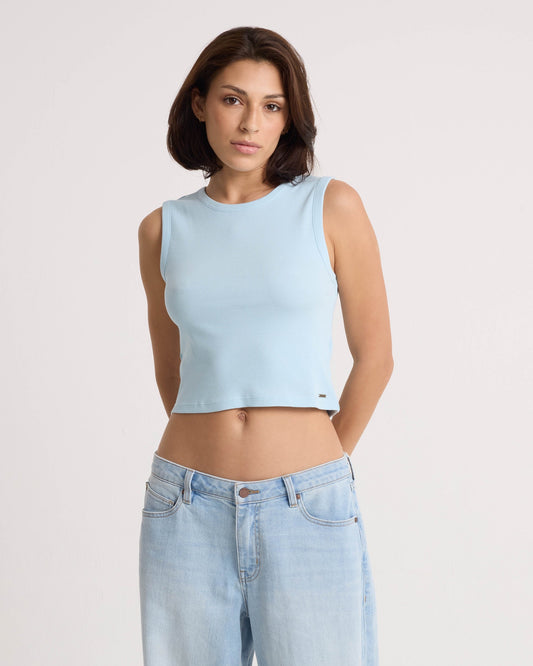 A person with short brown hair wears the Volcom Women's Mary Mack Tank in light blue and matching stretchy knit jeans. They stand against a plain white background, facing the camera with a neutral expression and hands behind their back.