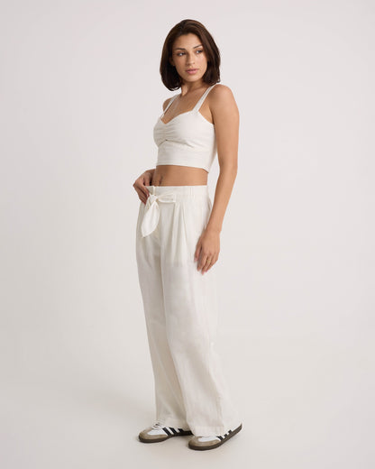 A woman models Volcom's Women's Bow Chicka Pants—loose, wide-leg, cotton-linen blend pants with a tie-front waist—paired with a white crop top and black and white sneakers against a plain background.