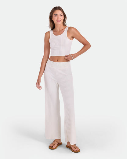 A woman smiles in a Volcom Women's Lounge Bra Tank paired with wide-leg pants and sandals, posing against a plain white background—an essential everyday outfit.