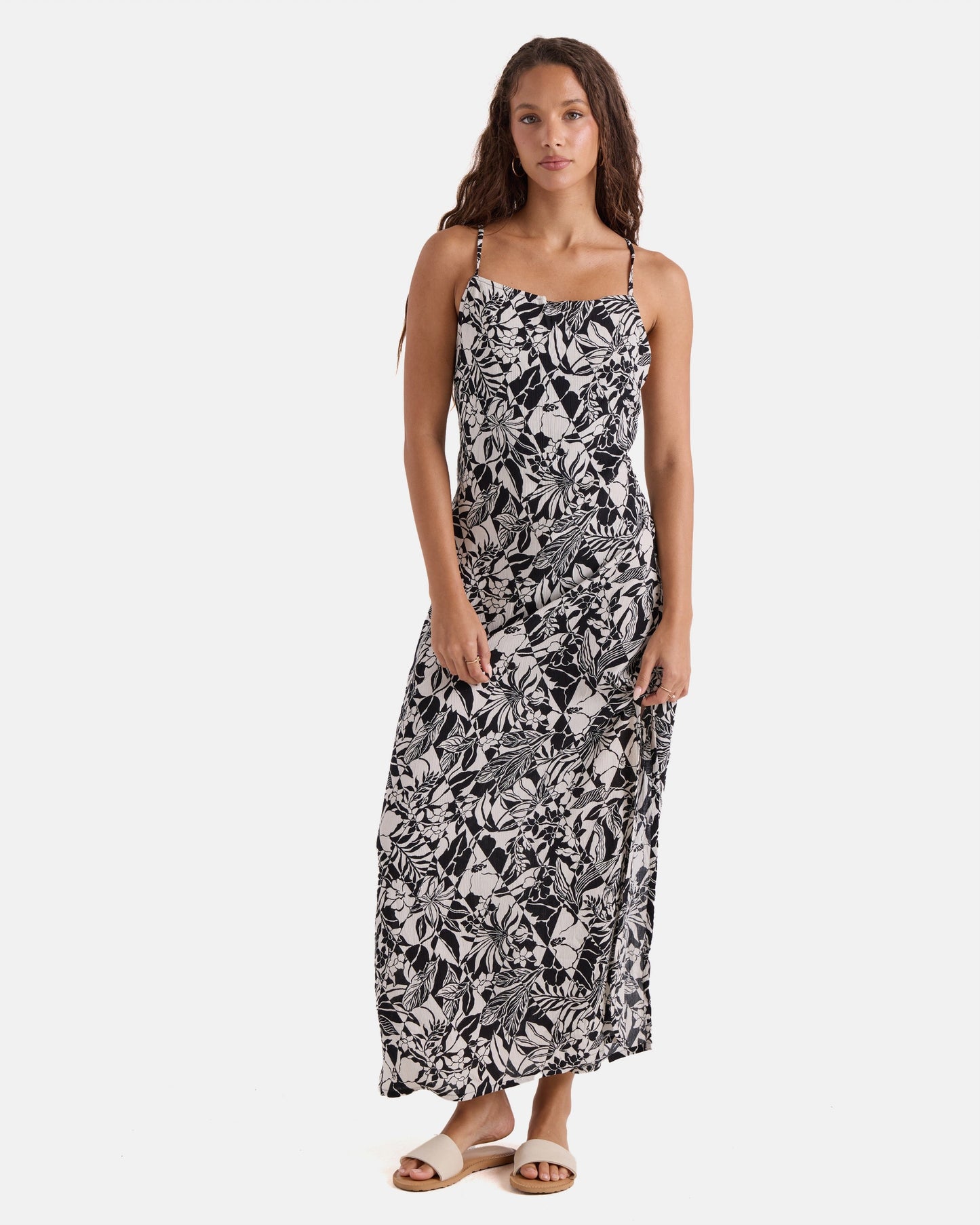 A woman with long curly hair wears the Volcom Women's Slip Maxi, a sleeveless black and white floral print dress with spaghetti straps and sandals, standing against a white background.