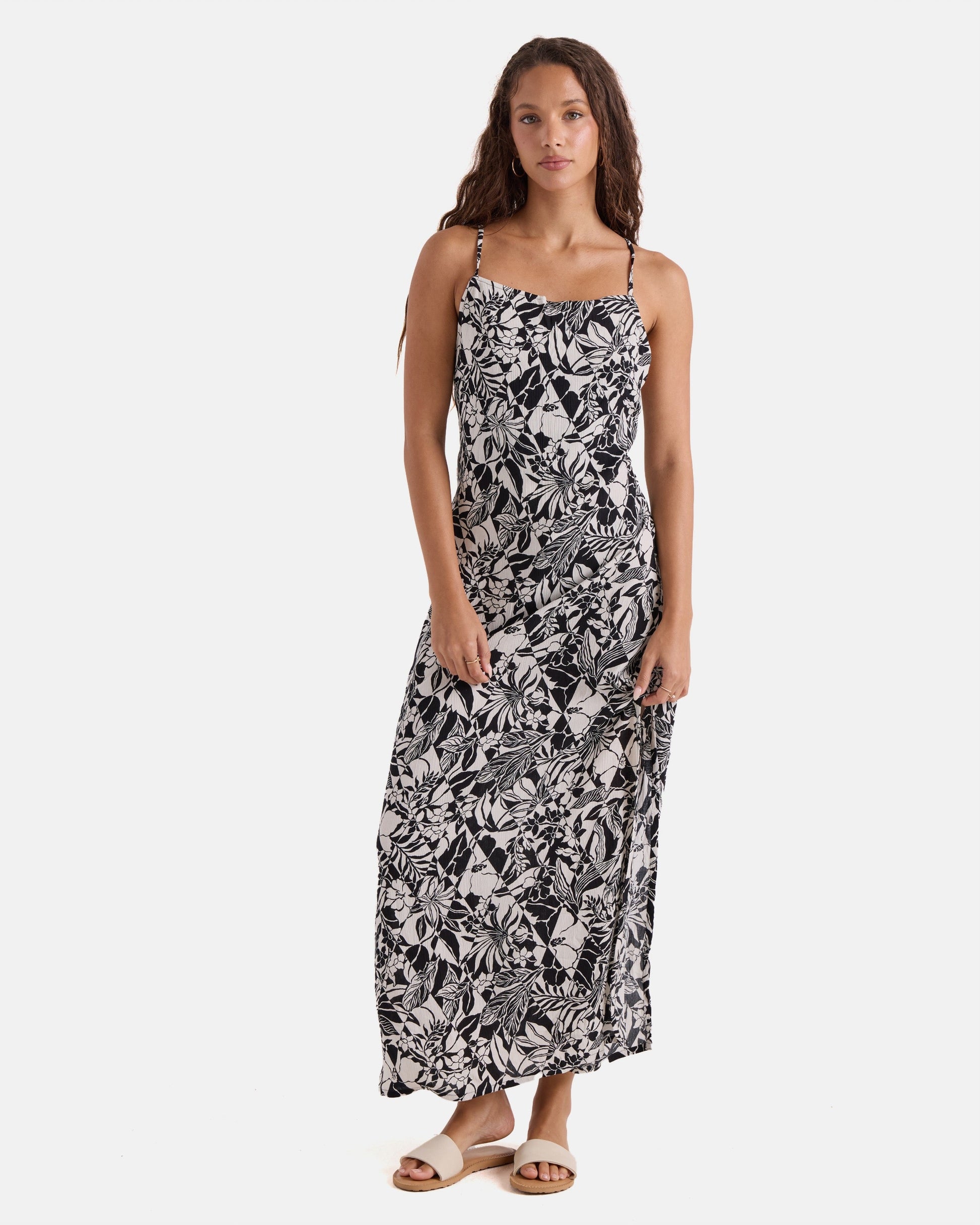 A woman with long curly hair wears the Volcom Women's Slip Maxi, a sleeveless black and white floral print dress with spaghetti straps and sandals, standing against a white background.
