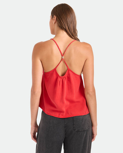 A woman with brown hair worn down faces away, wearing the Volcom Women's Pleat Heart Cami in Persimmon with crisscross straps and a relaxed fit, paired with dark gray pants against a plain white background.