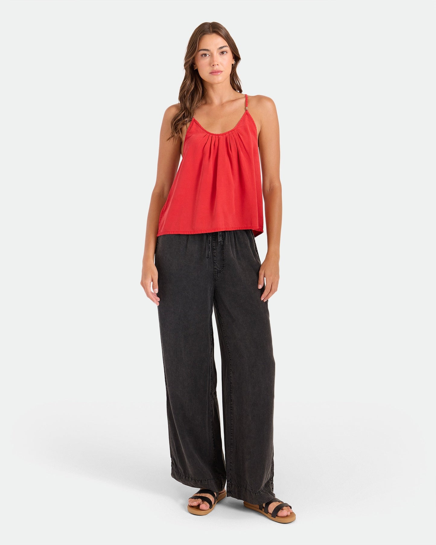 A woman with long hair wears the Volcom Women's Pleat Heart Cami in persimmon, paired with relaxed wide-leg black pants and black sandals, standing casually against a plain background.